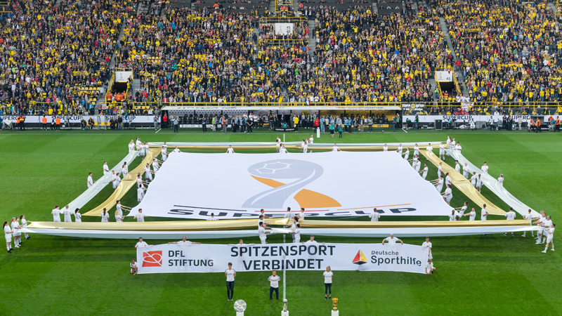 Auf dem Spielfeld des Dortmunder Stadions ist neben dem Supercup-Logo ein Banner mit der Aufschrift "Spitzensport verbindet" und den Logos von DFL Stiftung und Deutscher Sporthilfe zu sehen. Vor diesem Banner stehen drei Personen hinter den drei Trophäen des deutschen Fußballs, der Meisterschale, dem Supercup und dem DFB-Pokal.