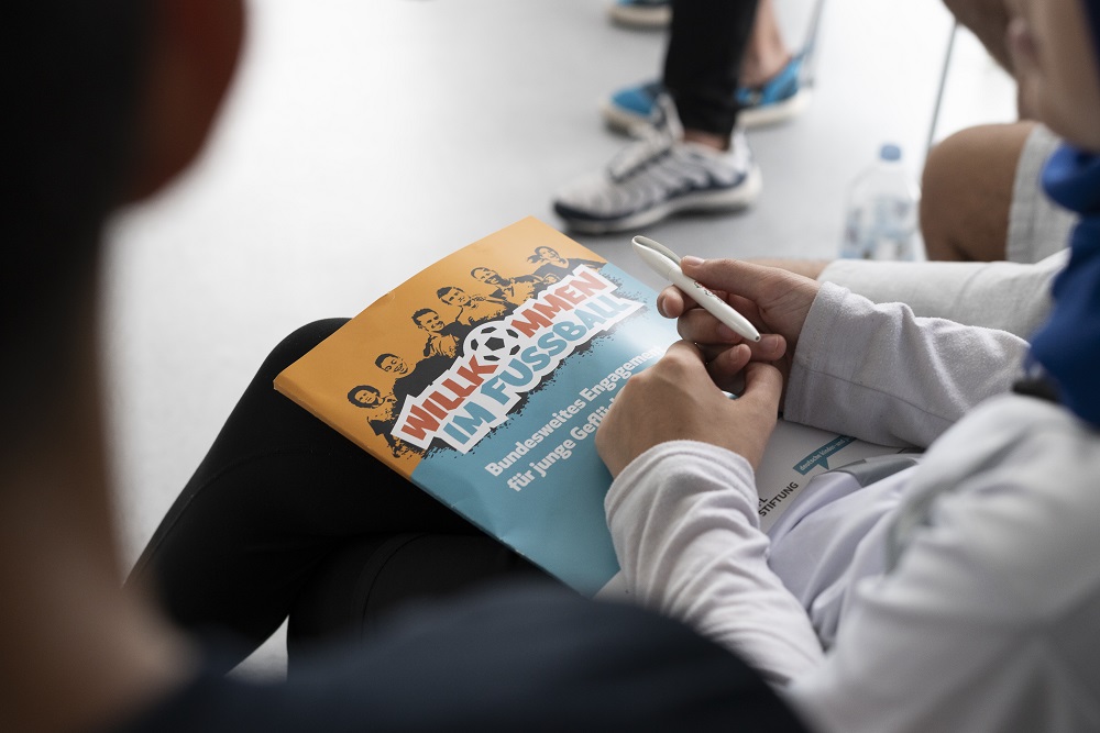 Ein Teilnehmende hält das Programmheft "Willkommen im Fußball" auf dem Schoß sowie einen Kugelschreiber in der Hand