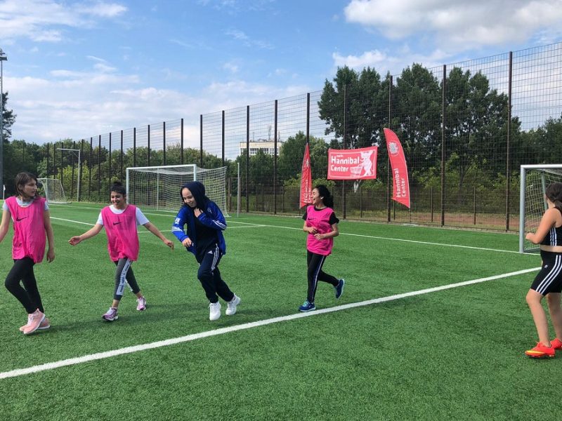Eine Gruppe von Jugendlichen spielt Fußball auf einem grünen Kunstrasenplatz. Einige Mädchen in Sportkleidung sind in Bewegung, während andere am Spielfeldrand stehen. Im Hintergrund sind Fußballtore und Bäume zu sehen.