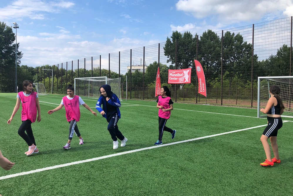 Eine Gruppe von Jugendlichen spielt Fußball auf einem grünen Kunstrasenplatz. Einige Mädchen in Sportkleidung sind in Bewegung, während andere am Spielfeldrand stehen. Im Hintergrund sind Fußballtore und Bäume zu sehen.
