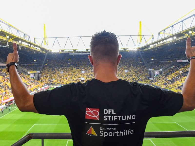Ein Mann im schwarzen T-Shirt mit Logos von der DFL Stiftung und Deutschen Sporthilfeguckt auf das Stadion und zeigt beide Daumen nach oben