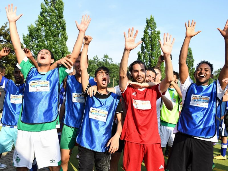 Junge Menschen mit erhobenen Händen stehen auf einem Fußballplatz und jubeln. Sie tragen Trainingsshirts in Blau und Rot und stehen im Fokus der Aufnahme. Im Hintergrund sind weitere Personen und Bäume zu sehen.