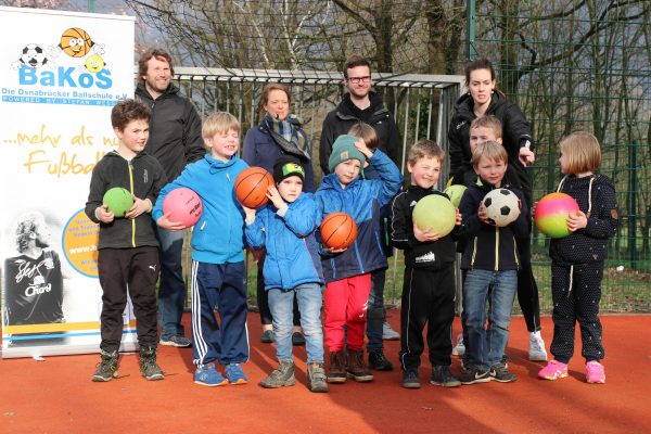 Eine Gruppe von Kindern steht auf einem roten Sportplatz und hält verschiedene Bälle hoch. Im Hintergrund steht ein Banner mit der Aufschrift 