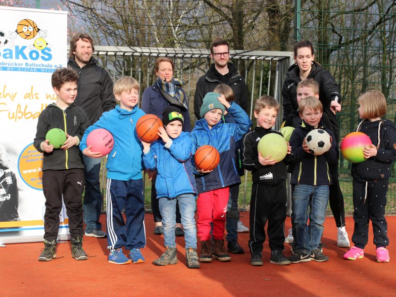 Eine Gruppe von Kindern steht auf einem roten Sportplatz und hält verschiedene Bälle hoch. Im Hintergrund steht ein Banner mit der Aufschrift 