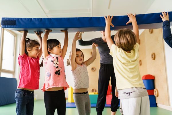 Mehrere Kinder tragen gemeinsam eine Turnmatte mit ausgestreckten Armen über ihren Köpfen.