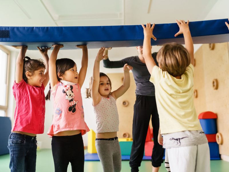 Mehrere Kinder tragen gemeinsam eine Turnmatte mit ausgestreckten Armen über ihren Köpfen.