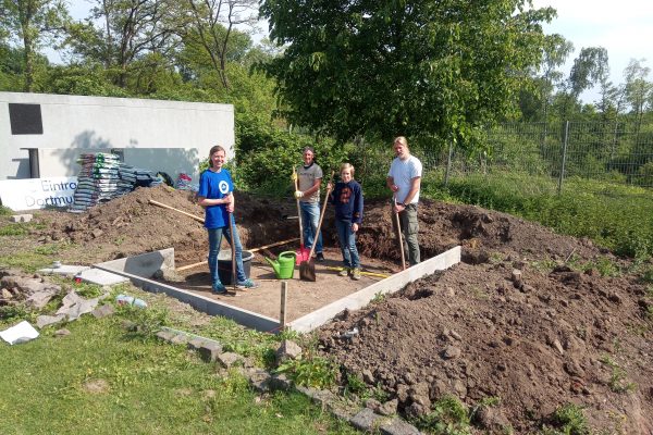 Vier Personen stehen mit Gartengeräten auf einem Fundament, auf dem ein Gewächshaus errichtet werden soll.