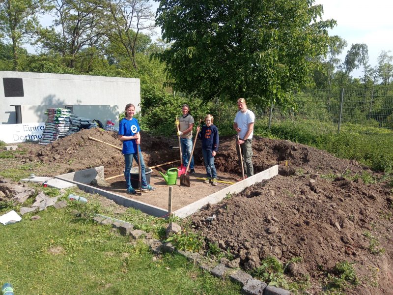 Vier Personen stehen mit Gartengeräten auf einem Fundament, auf dem ein Gewächshaus errichtet werden soll.