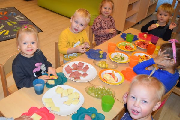 Mehrere Kleinkinder sitzen an einem mit bunten Tellern und verschiedenen gesunden Lebensmitteln gedeckten Tisch und lachen in die Kamera.