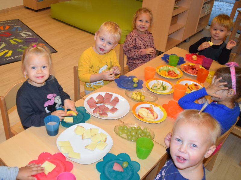 Mehrere Kleinkinder sitzen an einem mit bunten Tellern und verschiedenen gesunden Lebensmitteln gedeckten Tisch und lachen in die Kamera.