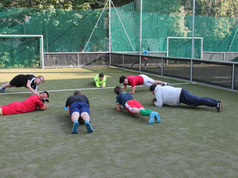 Sieben junge Menschen mit und ohne Behinderung machen gemeinsam auf einem Fußballplatz Kräftigungsübungen.