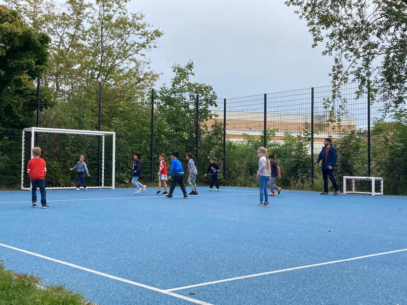 Mehrere Kinder spielen auf einem Bolzplatz mit einem blauen Bodenbelag gemeinsam Fußball.