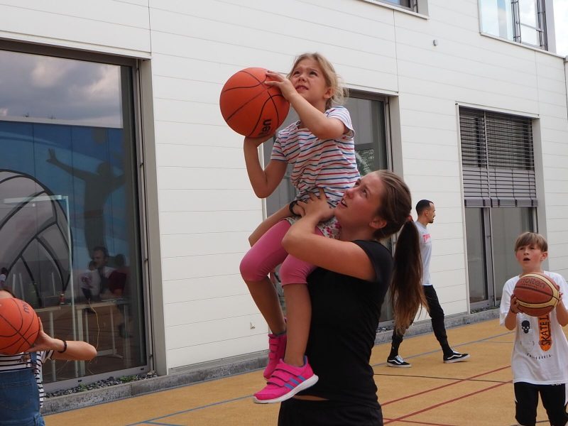 Ein Mädchen sitzt auf der Schulter einer jungen Frau und hält einen Basketball in der Hand. Die junge Frau hilft ihr dabei, den Ball in den Korb zu werfen.