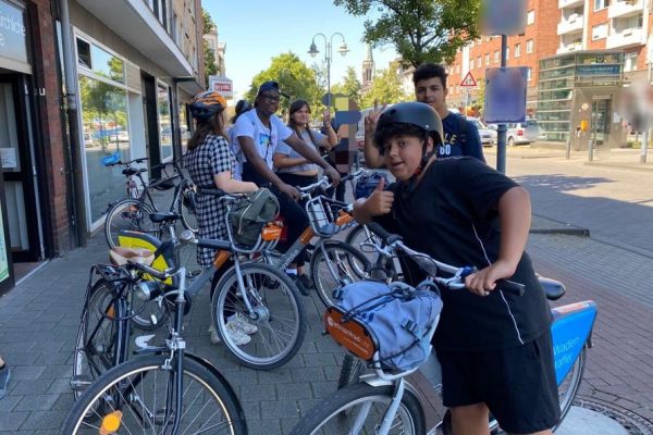 Mehrere Kinder stehen mit ihren Fahrrädern am Straßenrand und lachen in die Kamera.
