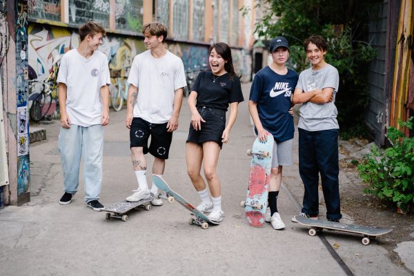 Fünf junge Menschen verschiedener Menschen stehen mit ihren Skateboards auf einer Straße und lachen in die Kamera.