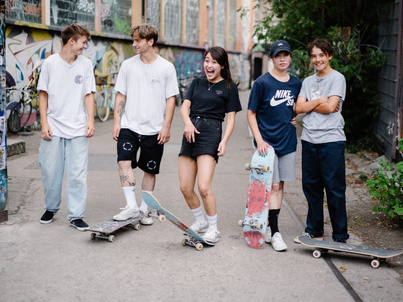 Fünf junge Menschen verschiedener Menschen stehen mit ihren Skateboards auf einer Straße und lachen in die Kamera.
