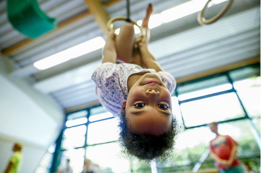Ein Mädchen hängt mit ihren Füßen in einem Ring, an dem sie sich auch noch mit den Händen festhält. Dabei schaut sie von oben auf die Kamera hinab.