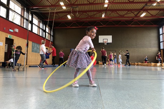 In einer Turnhalle steht ein kleines Mädchen, das mit beiden Händen einen gelben Hula-Hoop-Refen auf Höhe ihrer linken Hüfte greift. Der gegenüberliegende Punkt des Hula-Hoop-Reifens berührt dahingegen noch den Boden.lle den Boden berührt.