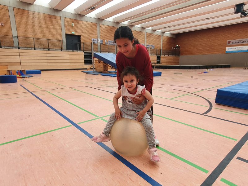 Kleines Mädchen sitzt auf dem Gymnastikball in der Turnhalle während sie ein älteres Mädchen hält