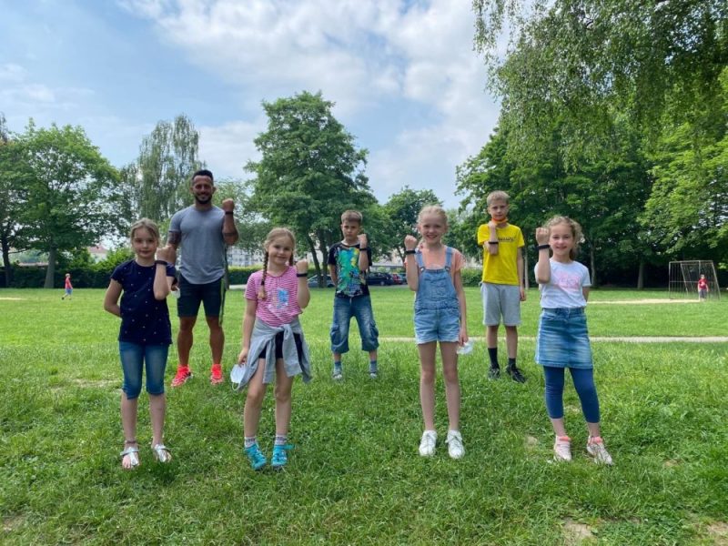 Sechs Schülerinnen und Schüler stehen mit ihrem Lehrer auf dem Fußballfeld und zeigen die Fitnessarmbänder