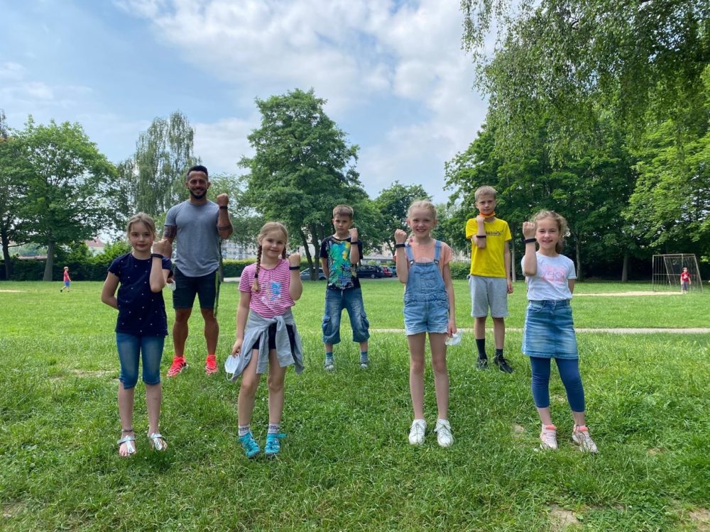 Sechs Schülerinnen und Schüler stehen mit ihrem Lehrer auf dem Fußballfeld und zeigen die Fitnessarmbänder