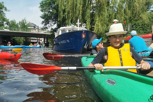 Ein Junge sitzt in einem grünen Kajak, das direkt am Flussufer steht. Er trägt eine gelbe Schwimmweste und einen hellen Hut. In seinen Händen hält der Junge ein Paddel. Weiter hinter im Kajak sitzt in einer weiteren Sitzluke ein anderer Junge mit Sonnenbrile und blauer Cappy. Auch er hält ein Paddel in der Hand. Im Hintergrund fahren weitere Kinder mit Kajaks auf dem Fluss, am Ufer steht ein Boot.