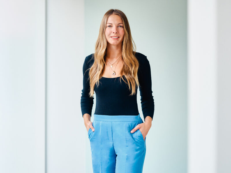 Andrea Bonk, lächelnde Frau mit langen, blonden Haaren, trägt ein schwarzes Langarmshirt und blaue Stoffhosen und steht selbstbewusst vor einem hellen Hintergrund.