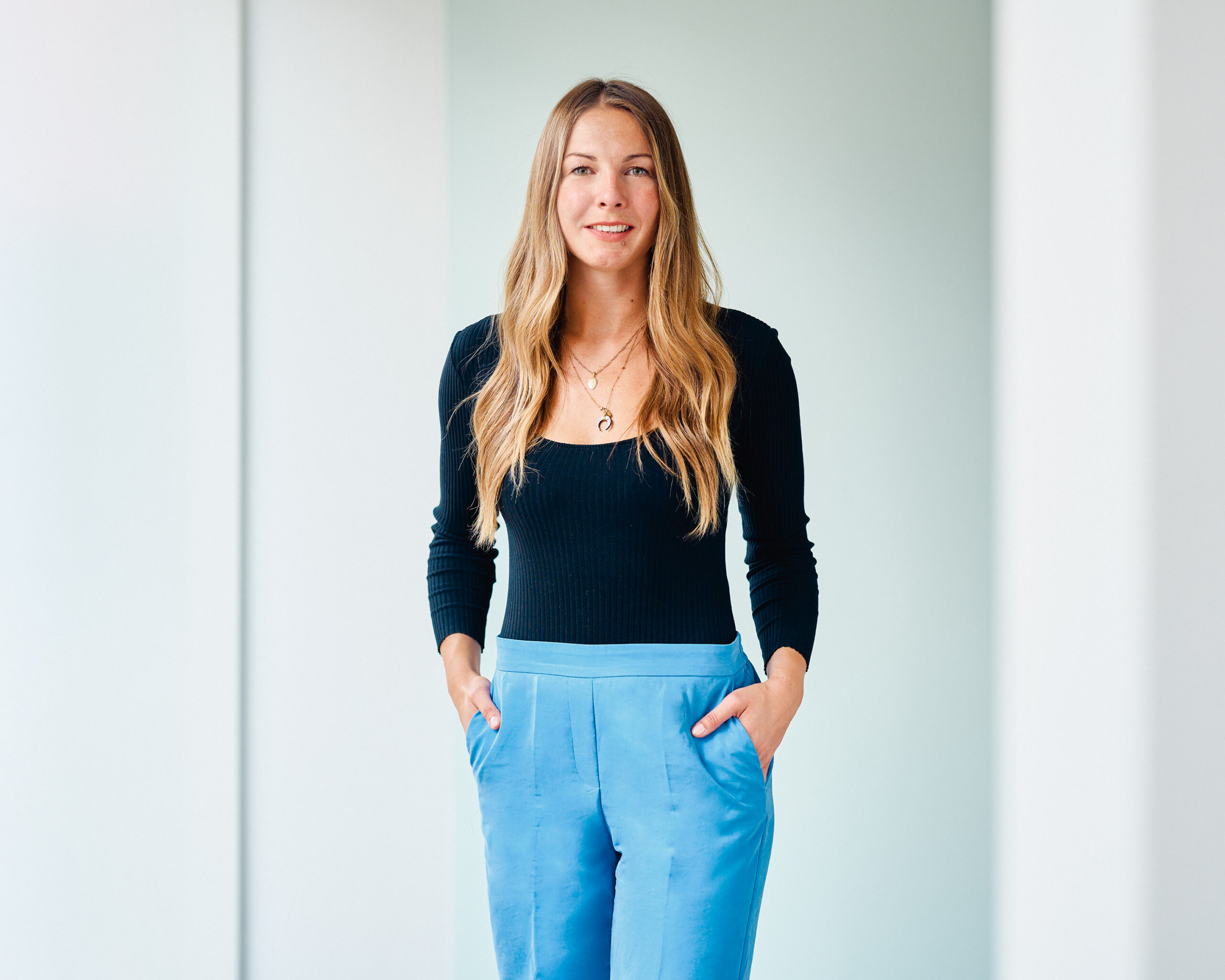 Andrea Bonk, lächelnde Frau mit langen, blonden Haaren, trägt ein schwarzes Langarmshirt und blaue Stoffhosen und steht selbstbewusst vor einem hellen Hintergrund.