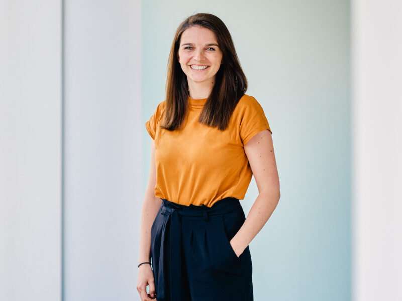Frederike Suckert, lächelnde Frau mit braunen Haaren und orangefarbenem Oberteil, posiert vor hellem Hintergrund.