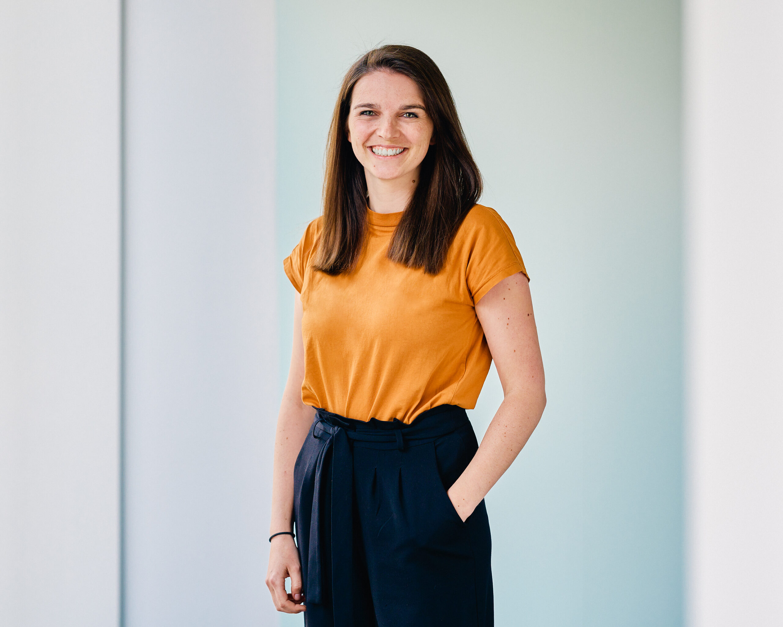 Frederike Suckert, lächelnde Frau mit braunen Haaren und orangefarbenem Oberteil, posiert vor hellem Hintergrund.