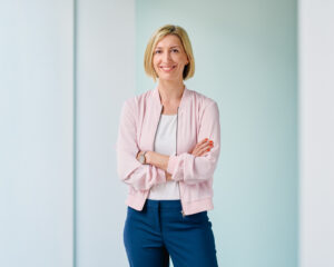 Franziska Fey, lächelnd mit verschränkten Armen vor hellblauem Hintergrund.