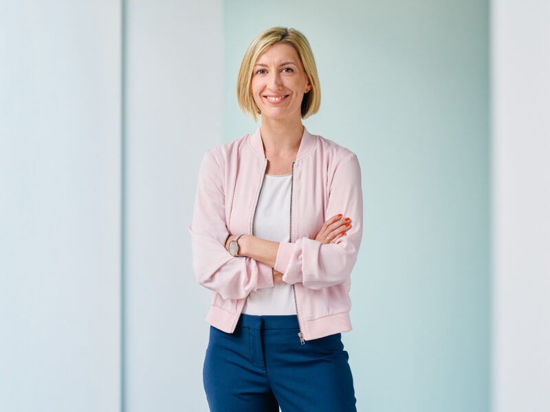 Franziska Fey, lächelnd mit verschränkten Armen vor hellblauem Hintergrund.