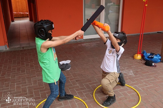Zwei Kinder stehen jeweils in einem mit einem Ring markierten Bereich und treten mit Schaumstoff-Schlägern gegeneinander an. Dabei tragen sie jeweils einen Kopfschutz.
