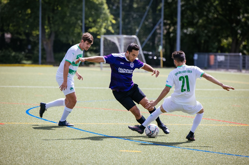 Ein Fußballspieler in blauem Trikot dribbelt den Ball während eines Spiels, während zwei Gegner in weißen Trikots ihn verfolgen. Im Hintergrund ist ein grüner Fußballplatz und Zuschauertribünen zu sehen.