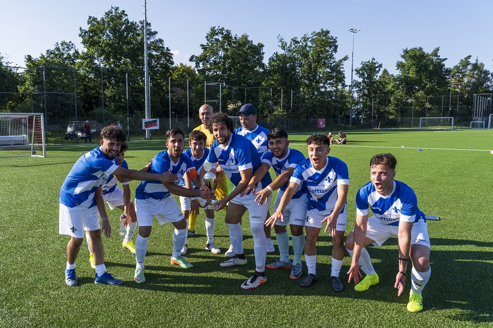 Eine Gruppe junger Fußballspieler in blauen Trikots und weißen Shorts steht auf einem grünen Fußballfeld und hält sich an den Händen, während sie gemeinsam jubeln. Im Hintergrund ist ein Fußballtor und grüne Bäume zu sehen.