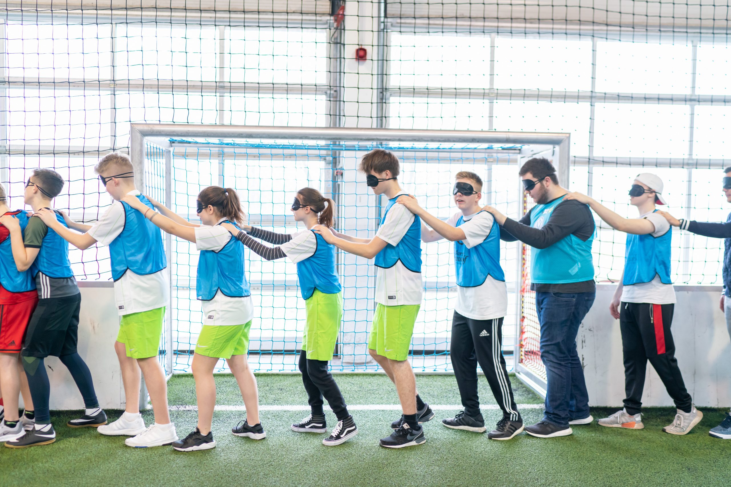 Eine Gruppe von Jugendlichen steht in einer Sporthalle in einer Reihe und hält sich an den Schultern fest. Alle tragen blaue Westen und einige haben verbundenen Augen, um sich gegenseitig zu führen. Im Hintergrund ist ein Fußballtor zu sehen.