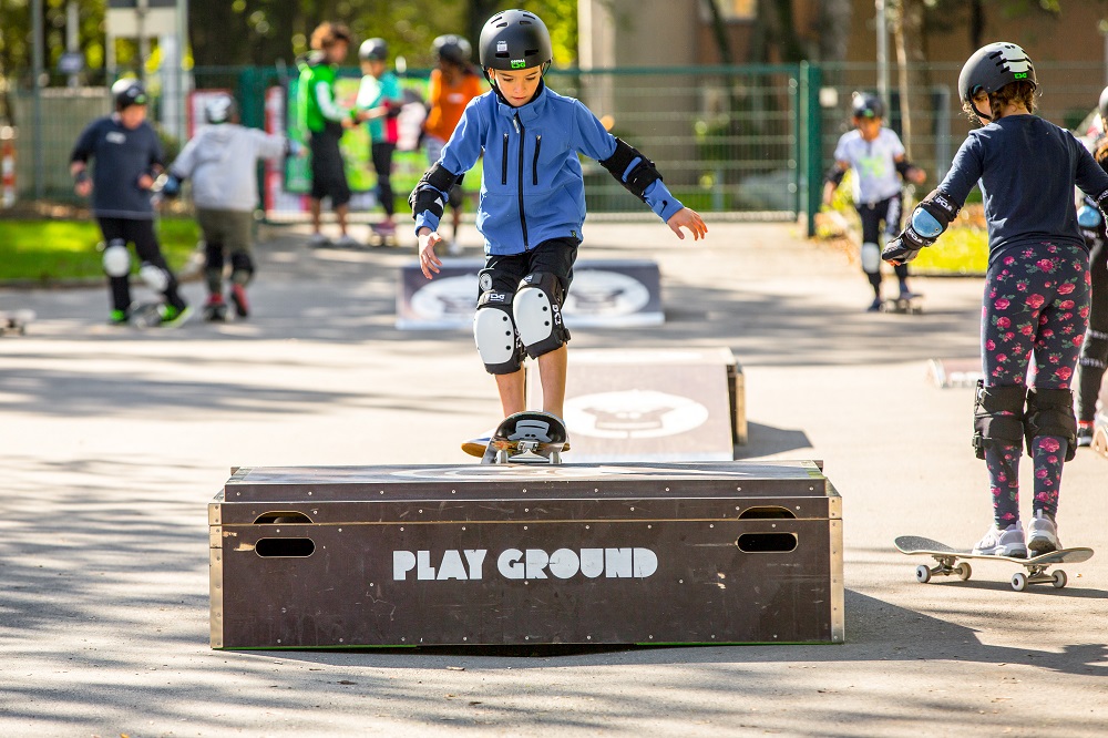 Ein Junge fährt auf einem Skateboard über eine erhöhte Plattform mit der Aufschrift "PLAY GROUND". Im Hintergrund sind weitere Kinder und Jugendliche auf Skateboards oder Fahrrädern zu sehen.