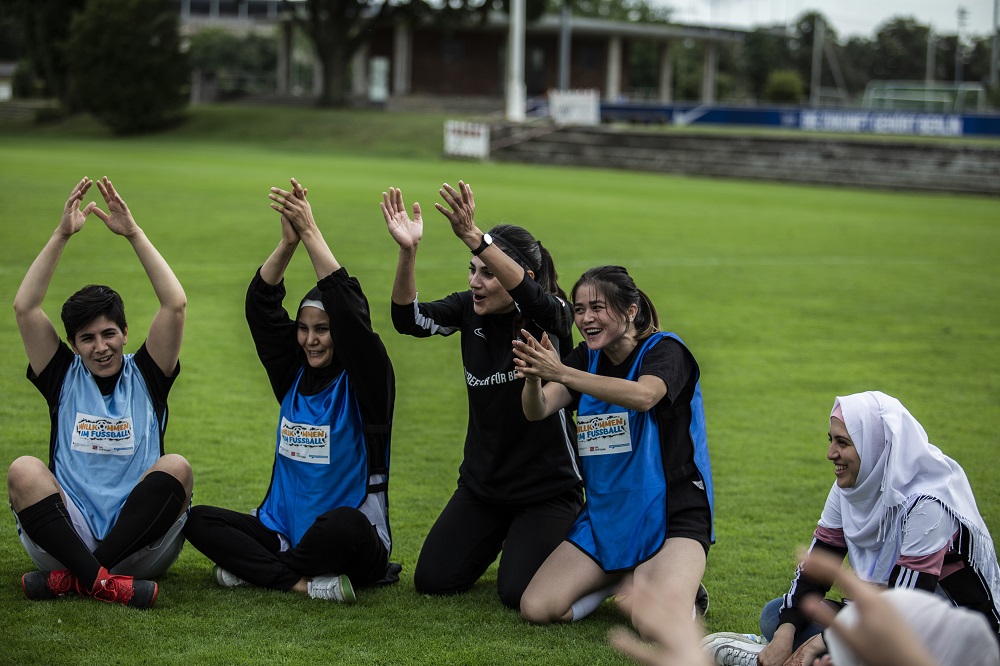 Eine Gruppe von fußballbegeisterten Frauen mit Kopftuch feiert auf einem grünen Rasen. Sie heben die Arme in die Luft und lächeln in die Kamera. Auf ihren Trainingswesten ist der Schriftzug "Willkommen im Fußball" zu sehen.
