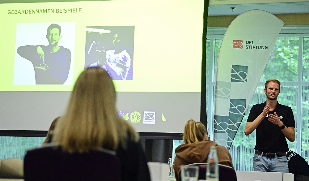 Florian Hansing vom DEAF BVB Fanclub e.V. erklärt vor einer Gruppe von Teilnehmenden auf einer Veranstaltung der DFL Stiftung die Grundlagen der Gebärdensprache, während auf einer Leinwand Beispiele für Gebärden zu sehen sind.