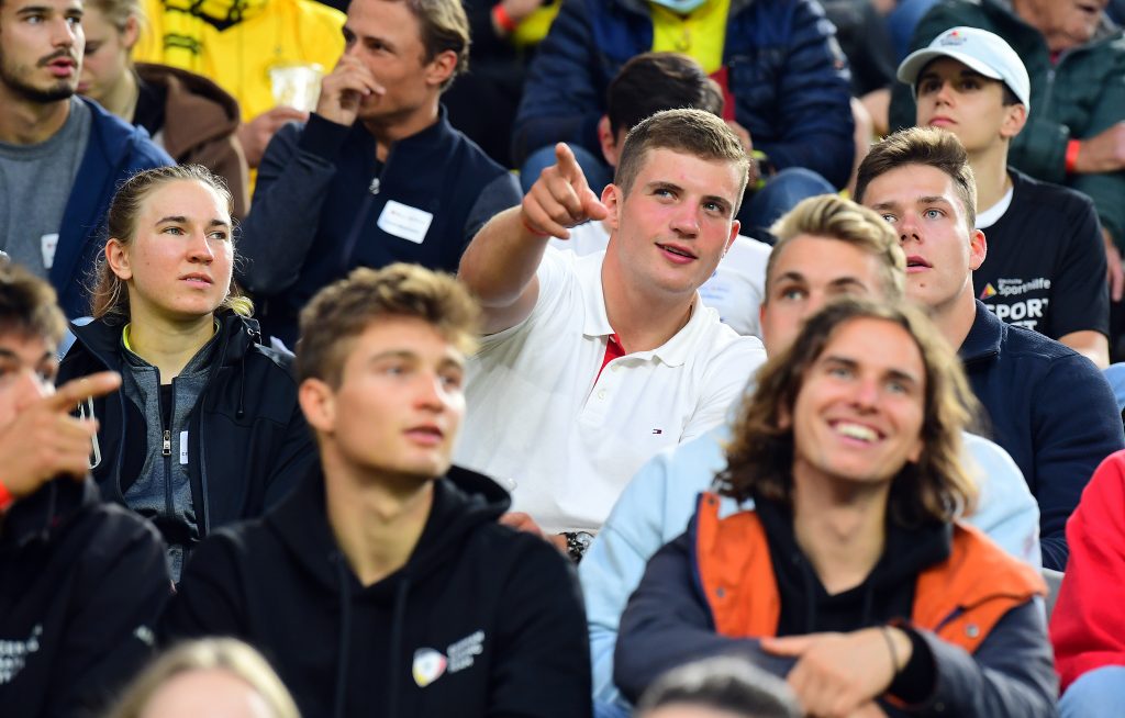 Young people are sitting in the stands at the soccer stadium. One of them points his finger at something.