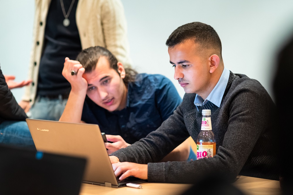 Zwei junge Männer sitzen an einem Tisch und arbeiten an einem Laptop. Der eine Mann ist vertieft in die Tastatur, der andere hält nachdenklich die Hand an die Stirn. Neben dem Laptop steht eine Flasche stilles Wasser.