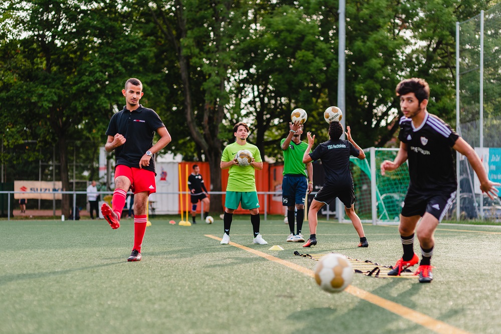 Eine Trainingsszene: Mehrere Jugendliche und ein Trainer stehen auf einem grünen Kunstrasenplatz und üben Fußball. Im Vordergrund ist ein Jugendlicher mit dem Ball am Fuß zu sehen, während ein Trainer Anweisungen gibt und andere Jugendliche im Hintergrund aufmerksam beobachten.