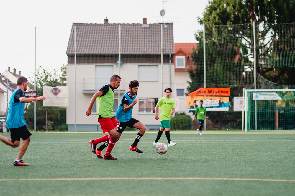 Fußballspieler in Trainingskleidung auf einem grünen Kunstrasenplatz. Im Hintergrund ein Wohnhaus und ein Zaun.