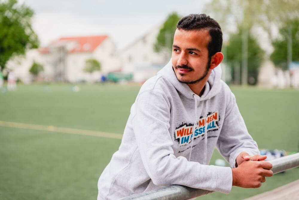 Junger Mann mit dunklen Haaren und grauem Hoodie lehnt lächelnd an einem Geländer, im Hintergrund ein Fußballfeld.