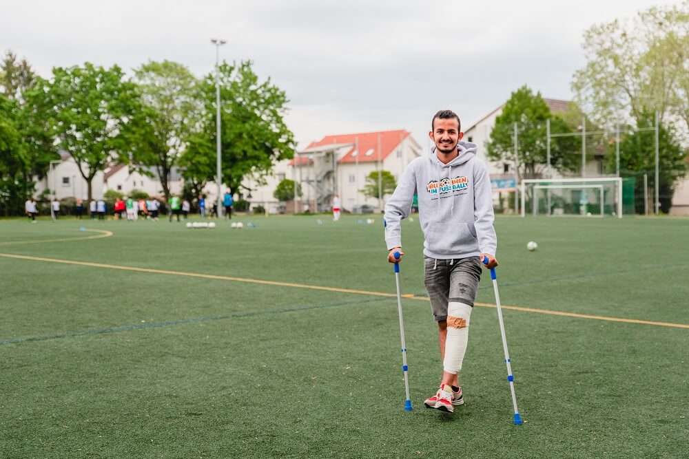 Ein junger Mann mit Gehstützen steht auf einem Fußballfeld. Er trägt einen grauen Hoodie, eine dunkle Hose und rote Turnschuhe. Im Hintergrund sind andere Personen und ein Vereinsgebäude zu erkennen.