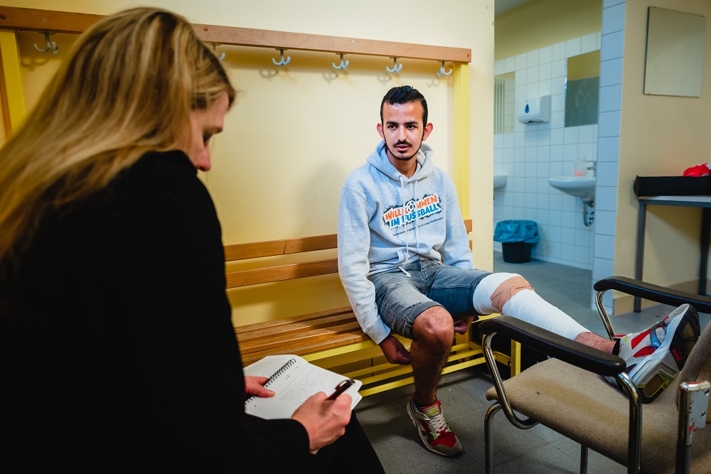 Ein Mann sitzt auf einer Umkleidebank, sein Bein ist in einer Schiene fixiert. Eine Frau sitzt ihm gegenüber und notiert etwas in einen Block. Im Hintergrund sind die spärlich eingerichteten Umkleidekabinen zu sehen.