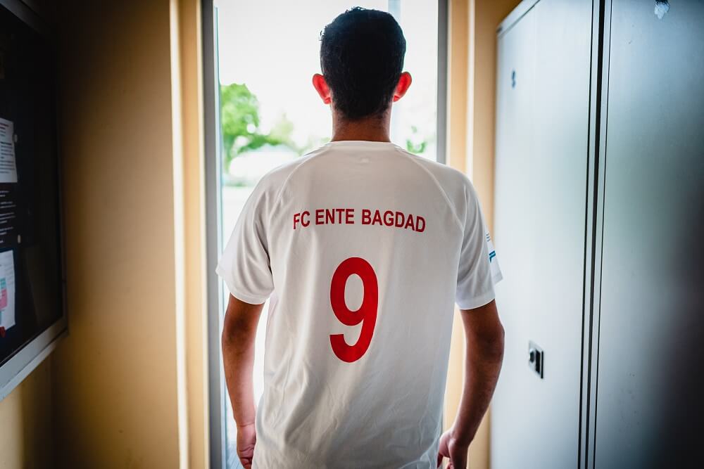 Ein Fußballspieler steht mit dem Rücken zum Betrachter da und trägt ein weißes Trikot mit der Aufschrift "FC ENTE BAGDAD" und der Rückennummer 9.