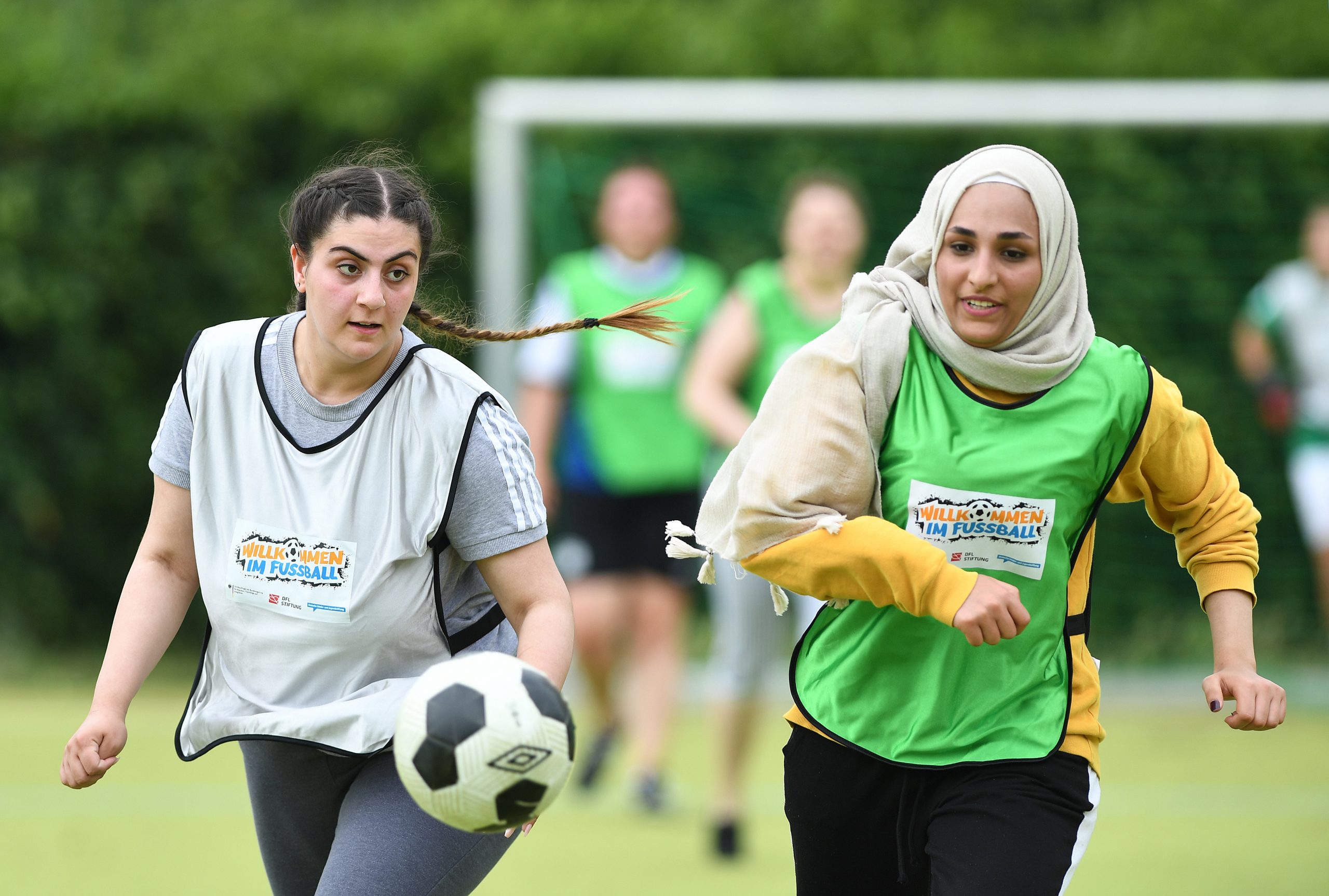 Zwei junge Frauen mit Kopftüchern und in Trainingsanzügen mit dem Aufdruck "Willkommen im Fußball" spielen Fußball auf einem grünen Rasenplatz. Die eine Frau schießt gerade den Ball, die andere rennt daneben. Im Hintergrund sind weitere Spielerinnen erkennbar.