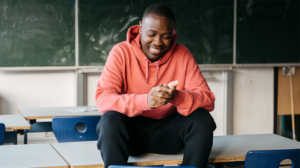 Matondo Castlo sitzt in einer Schulklasse auf einem Tisch. Im Hintergrund ist eine Tafel zu sehen.