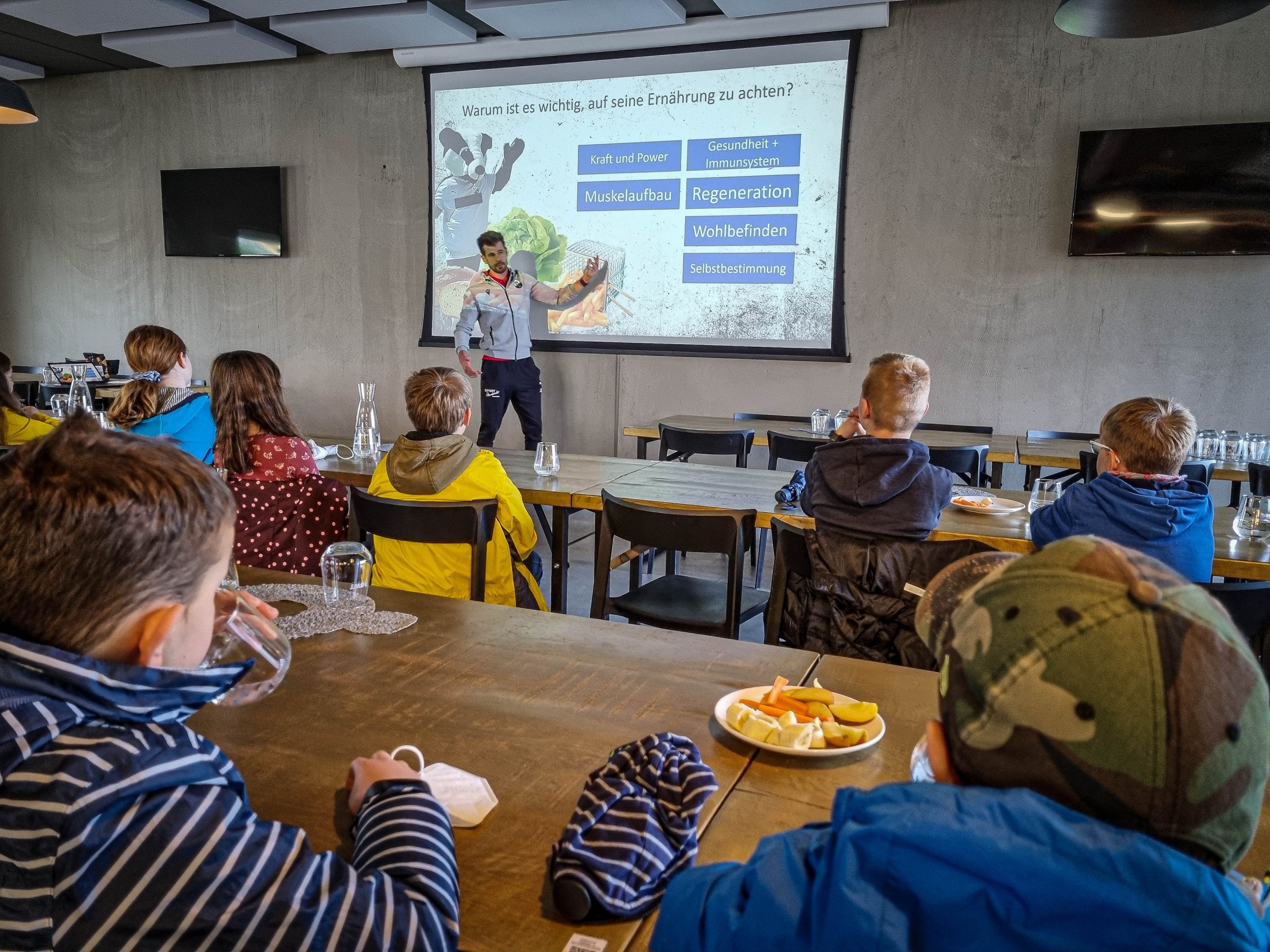 Eine Gruppe Jugendlicher in Fußballtrikots posiert auf einem grünen Kunstrasenplatz mit mehreren Erwachsenen. Im Hintergrund sind Bäume und ein Fußballfeld zu sehen.